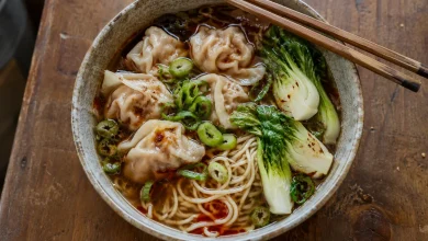Top-down view of wonton noodle soup in a ceramic bowl with dark beef broth, red chili oil, pleated pork wontons, bok choy, noodles and scallions with chopsticks