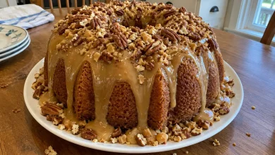 Whole caramel brown sugar pound cake on a white cake plate with caramel glaze dripping down the sides and loaded with chopped pecans and toffee bits