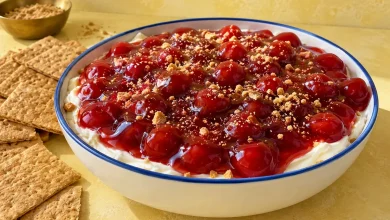 No-bake cherry cheesecake dip topped with glossy cherry pie filling in a round blue-rimmed stoneware dish with graham crackers on a butter-yellow plaster backdrop