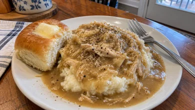 Plate of crockpot chicken and gravy over roasted garlic mashed potatoes with black pepper and a sweet dinner roll
