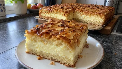 Big square slice of German bee sting cake on a plate showing caramelized almond top and thick cream layer, rest of sheet cake behind