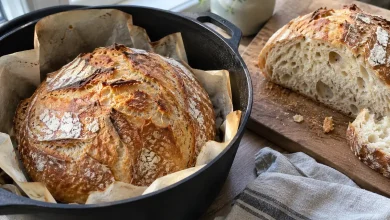 Golden crusty no knead bread loaf in an open Dutch oven on parchment with a cut half showing the airy crumb beside it