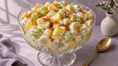 Creamy caramel fruit salad with bananas, pineapple and green grapes in a footed glass bowl with a caramel drizzle on a lavender plaster backdrop