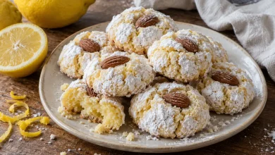 Plate of plump lemon almond cookies dusted with powdered sugar, each topped with a whole almond, one broken open to show the soft center, with fresh lemons nearby