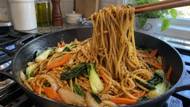 Dark skillet of glossy vegetable lo mein with chopsticks lifting a bundle of noodles, bok choy, carrots and onions visible