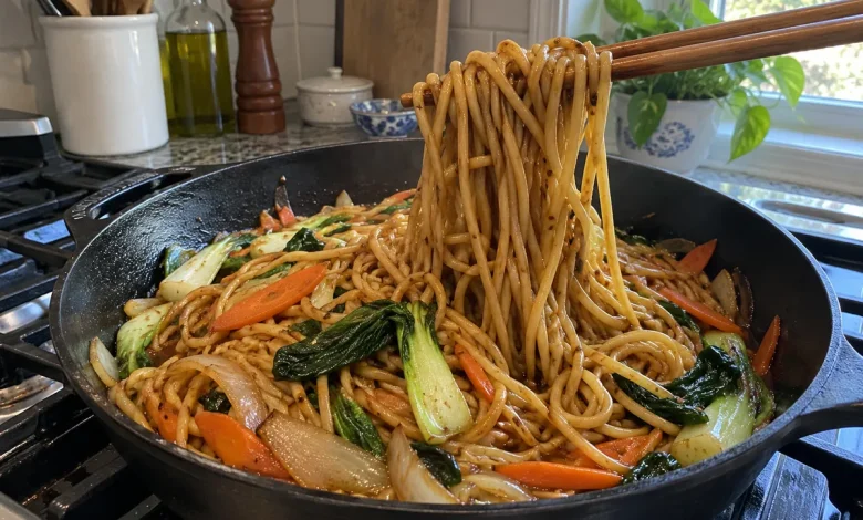Dark skillet of glossy vegetable lo mein with chopsticks lifting a bundle of noodles, bok choy, carrots and onions visible